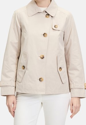 Vrouw draagt een beige jasje met grote knopen, klepzakken en verstelbare polsriempjes, gecombineerd met een witte broek.