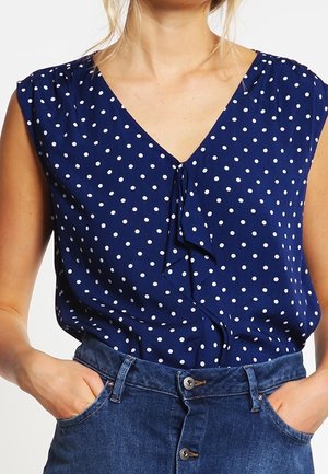 Kobieta w granatowej bluzce bez rękawów w białe grochy, włożonej do wysokich dżinsów z niebieskiego denimu, z rękami w kieszeniach.