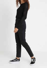 Kobieta ubrana w czarny długi sweter z koronkowymi rękawami, czarne wysokie dżinsy oraz czarno-białe trampki, stojąca na tle białym.