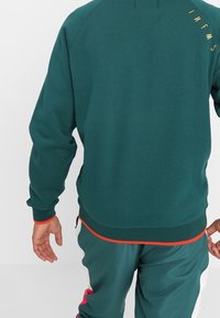 Turkusowa bluza z ribbowanymi mankietami i dołem, posiada złote logo na plecach. Noszona z pasującymi ciemnozielonymi dresami i czerwonymi akcentami.