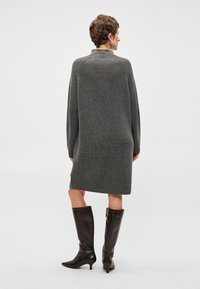 Abito grigio a maglia con colletto alto e maniche lunghe, abbinato a stivali neri di pelle alti fino al ginocchio e un design elegante. Finitura texturizzata visibile.