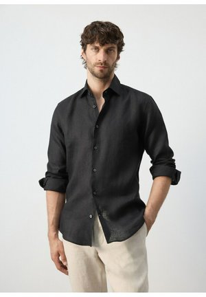 Hombre con cabello rizado que lleva una camisa negra de manga larga abotonada con las mangas remangadas y pantalones de color claro, de pie con una mano en el bolsillo.