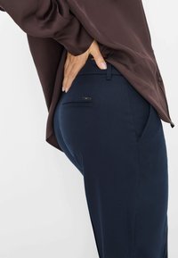 Marineblauwe broek met een gladde textuur en een slanke pasvorm, voorzien van een klein achterzakje met zilveren hardware en een bruine blouse die gedeeltelijk zichtbaar is.