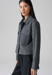 Grijze, kortgeknipte jas van textuurstof met een krijtstreep patroon, met twee borstzakken en een puntige kraag.