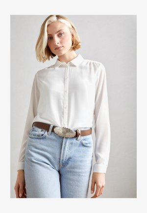 Hvid langærmet skjorte med knapper og en glat tekstur, parret med lyseblå højtaljede jeans og et brunt bælte med en sølv dekoreret spænde.