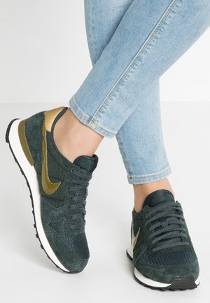 Persona con zapatillas Nike verde oscuro con detalles dorados y vaqueros ajustados azul claro, de pie sobre un suelo blanco.