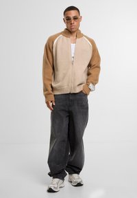 Beige und brauner Reißverschluss-Pullover mit gerippten Bündchen und Saum, der eine Mischung aus Texturen aufweist; getragen mit grauen, weiten Jeans und Turnschuhen.