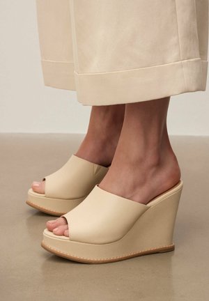 Bézs nyitott orrú ék szandál krémszínű bőszárú nadrággal viselve, sima padlón látható bokák és lábak.