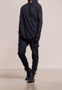 Musta huppari, valmistettu pehmeästä kankaasta, jossa pitkät hihat ja rento istuvuus. Yhdistetty mustien cargo-housujen kanssa, joissa on sivutaskut, ja tummien saappaiden kanssa.