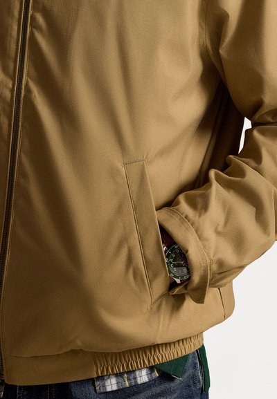 Brown zippered jacket made of smooth fabric, featuring a side pocket with a wristwatch partially visible, elasticized hem, and textured sleeves.