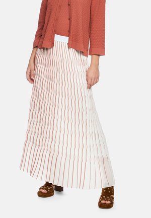 Frau trägt eine rostfarbene, strukturierte Strickjacke und einen weißen Faltenrock mit rostfarbenen vertikalen Wellenstreifen, kombiniert mit braunen Plateau-Sandalen.