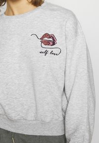 Sudadera gris con un gráfico de labios de lentejuelas en rosa y rojo, con el texto "amor propio" debajo. La tela parece suave y texturizada.