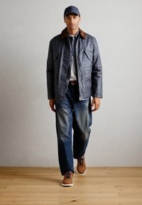 Graue Jacke mit braunem Kragen, weißes Hemd und blaue Jeans. Braune Schuhe mit weißen Sohlen. Das Model steht auf einem Holzboden vor einem hellen Hintergrund.