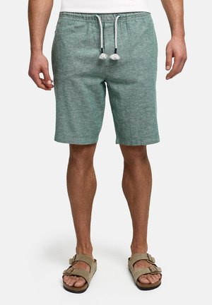 Man draagt groene trekkoortshorts en beige sandalen met gesp, staand tegen een effen witte achtergrond.