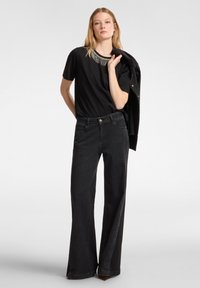 Camiseta negra con detalle de flecos decorativos en el escote, combinada con jeans negros de cintura alta y pierna ancha. Tejido texturizado, corte casual.