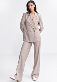 Beżowa dwurzędowa marynarka z brązowymi guzikami, w zestawie z dopasowanymi spodniami o szerokich nogawkach, ozdobionymi cienkimi pionowymi paskami i gładką fakturą.
