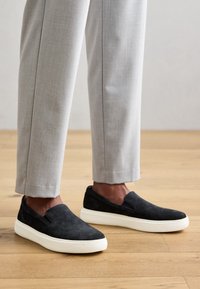 Scarpe slip-on in suede nere con suole in gomma bianche, abbinate a pantaloni su misura grigio chiaro. Sfondo in legno.
