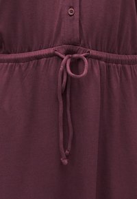 Κοντινό πλάνο σε μπορντό υφασμάτινο φόρεμα με δέσιμο στη μέση και εμφανές κουμπί πάνω από τη μέση.