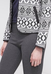 Gros plan d'une personne portant un pantalon gris ajusté et une veste à motif géométrique noir et blanc avec une poche bordée de noir.