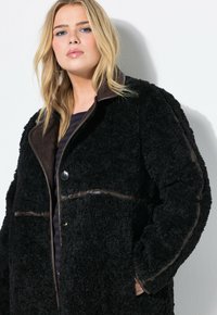 Giacca in pelliccia sintetica nera con dettagli in pelle marrone, grandi bottoni e colletto ampio. Tessuto testurizzato e vestibilità ampia e oversize.