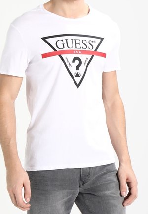 Hvid T-shirt med sort trekantet GUESS-logo, rød stribe og teksten "U.S.A.", båret af en person iført grå jeans, synlig fra taljen og ned.