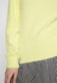 Puszysty żółty sweter z prążkowanymi mankietami i krawędzią; zestawiony z czarno-białą tkaniną w kratę psich zębów. Miękka tekstura i żywy kolor.