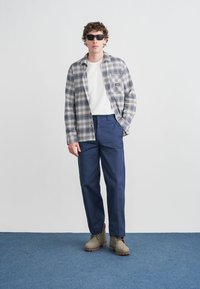 Καρό πουκάμισο σε μπλε και λευκό, λευκό μπλουζάκι από μέσα, ναυτικό παντελόνι και μπεζ μπότες, φορεμένα με γυαλιά ηλίου, πάνω σε φωτεινό φόντο.