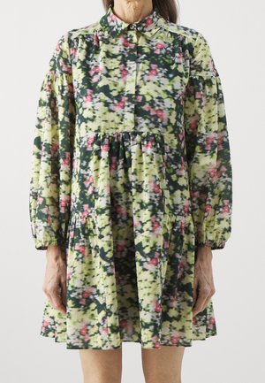 Femme portant une robe fleurie ample avec de longues manches bouffantes et un col boutonné aux couleurs vert, rose et blanc.
