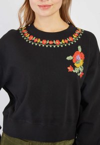 Kobieta w czarnej bluzie z kolorowymi haftowanymi wzorami kwiatowymi wokół dekoltu i w górnej lewej części klatki piersiowej.