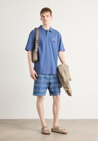 Blå pikétröja med en bröstlogotyp, parat med blå rutiga shorts. Brun budväska och beige jacka, bärs med bruna sandaler.