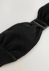 Svart texturerad hårband med ett vävt mönster, med ett elastiskt mittstycke och en rektangulär plastklämma för säker fastsättning.