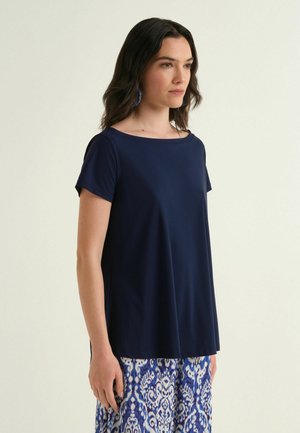 Donna con capelli scuri che indossa una maglietta blu navy a maniche corte e pantaloni blu con motivo, in piedi davanti a uno sfondo chiaro uniforme.