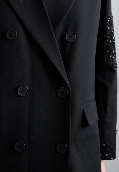 Melna divrindu veste ar divām teksturētām pogām, vienu slīpu kabatu un melno sekvīnu rotājumu uz piedurknes.
