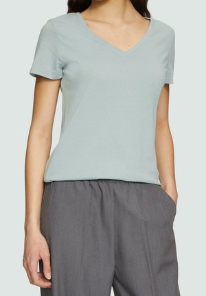 Frau trägt ein hellblaues kurzärmeliges V-Ausschnitt-T-Shirt und lockere graue Hosen, Hände in den Taschen, steht vor einfachem Hintergrund.