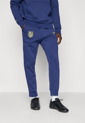 Mężczyzna w niebieskich dresowych spodniach Nike i dopasowanej bluzy z logo Atletico Madryt, czarnych sneakersach i białych skarpetkach, stojący na jednolitym tle.