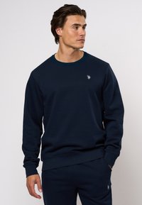 Námornícky modrá mikina vyrobená z mäkkého materiálu, s okrúhlym výstrihom a ribbenými manžetami. Vyšívané logo na hrudi, minimalistický dizajn.
