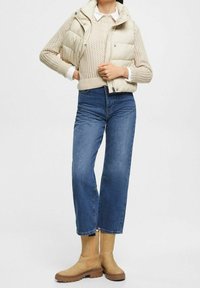 Beige Steppweste über einem cremefarbenen Strickpullover, kombiniert mit blauen geraden Jeans und hellbraunen Stiefeletten mit gerillter Sohle.