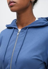 Μπλε φούτερ με φερμουάρ, κατασκευασμένο από μαλακό ύφασμα, διαθέτει κουκούλα με κορδόνι, ασημένιο φερμουάρ και λεία υφή. Δεν υπάρχουν ορατά σχέδια ή διακοσμητικά στοιχεία.