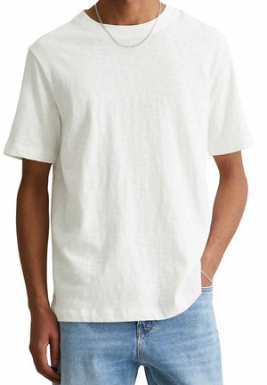 Homme portant un t-shirt blanc uni manches courtes, jean bleu clair, collier chaîne argentée et bracelet argenté, main dans la poche, torse visible.