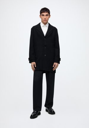 Jeune homme portant un manteau noir boutonné, un pantalon noir, une chemise blanche et des chaussures habillées noires, debout devant un fond blanc uni.
