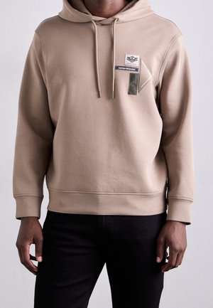 Mężczyzna w beżowej bluzie z kapturem z troczkami i trzema małymi naszywkami z logo Emporio Armani na piersi, w połączeniu z czarnymi spodniami.