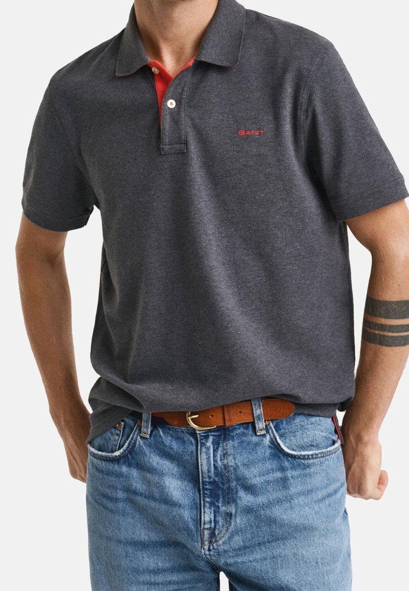 Homme portant un polo gris foncé avec un détail de col rouge et un logo, un jean bleu clair, une ceinture marron et un tatouage au bras avec des bandes noires.
