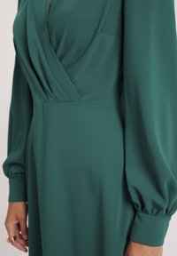 Zielona, długa sukienka z długimi rękawami, głębokim dekoltem w kształcie litery V oraz plisowanym gorsetem. Posiada zgromadzone bufiaste rękawy oraz gładki, teksturowany materiał.
