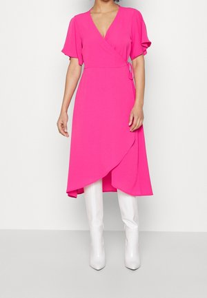 Femme portant une robe portefeuille rose vif à manches évasées et des bottes blanches montantes jusqu'aux genoux, debout sur un fond blanc.