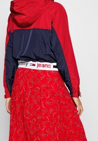 Röd och marinblå vindjacka med huva, tillsammans med en röd kjol med paisleymönster och en vit midja märkt "Tommy jeans."