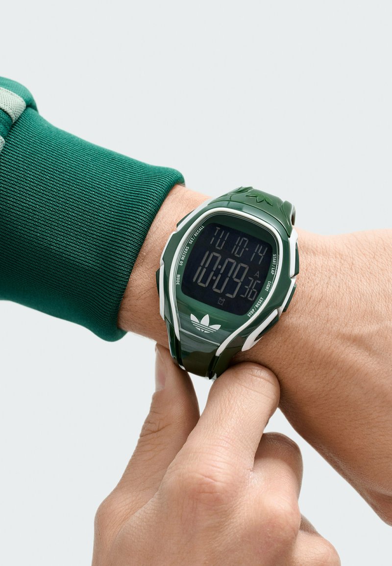 Reloj deportivo digital con correa de silicona verde, pantalla digital negra y detalles en blanco. Cuenta con funciones de hora, fecha y cronómetro.