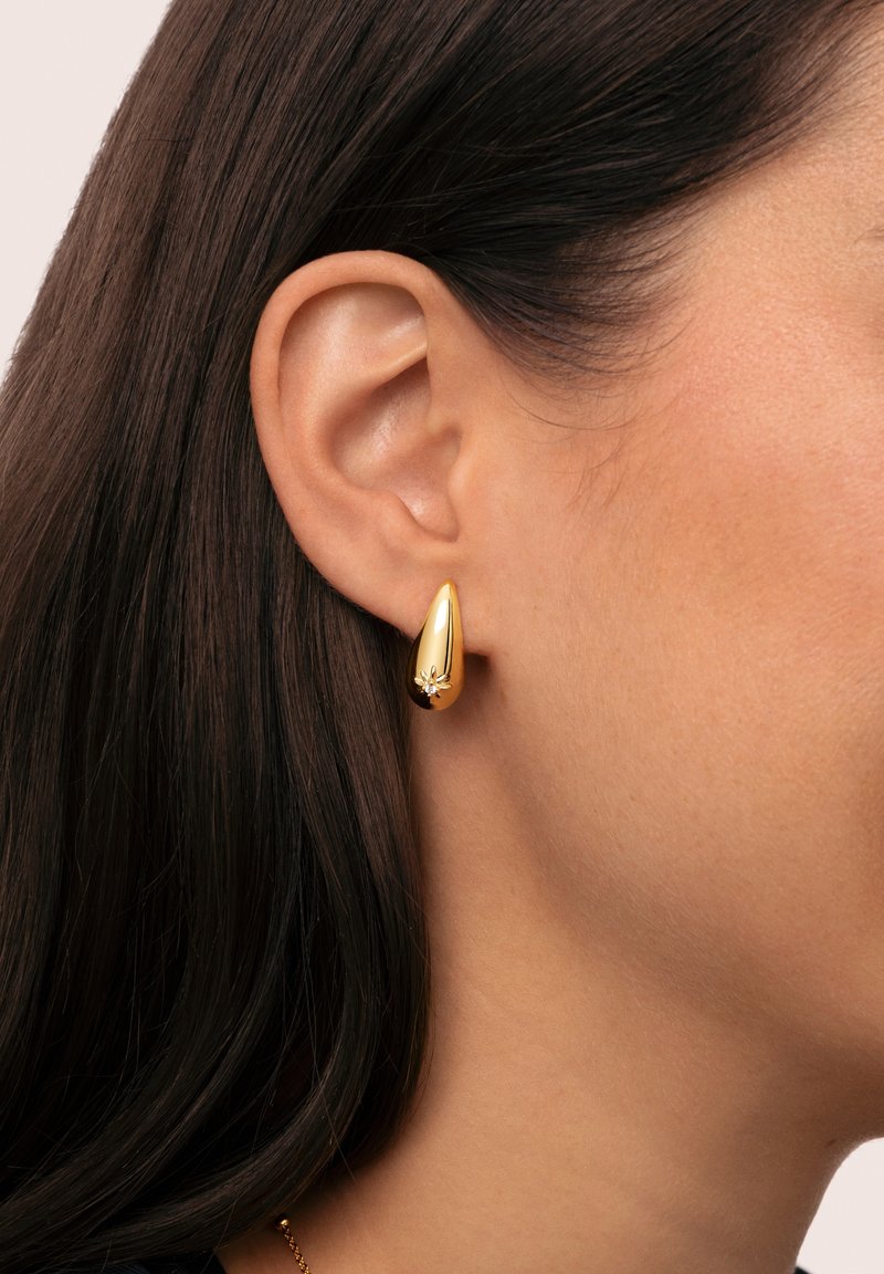 Pendientes en forma de lágrima de oro con un acabado pulido y un pequeño acento decorativo, exhibidos en la oreja de un modelo contra un cabello oscuro.