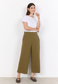 Woman with long dark hair wearing a white short-sleeve shirt, olive wide-leg cropped pants, and dark studded flats standing against white wall.