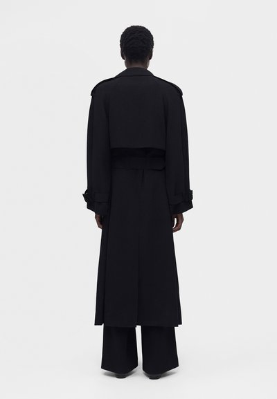 Άτομο με κοντά σγουρά μαλλιά φορά ένα μακρύ μαύρο παλτό και φαρδιά παντελόνια, στέκεται με την πλάτη σε μια απλή ανοιχτή φόντο.