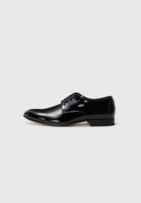 Zapato de vestir para hombre de piel negra pulida con cordones y tacón bajo, sobre un fondo gris claro liso.